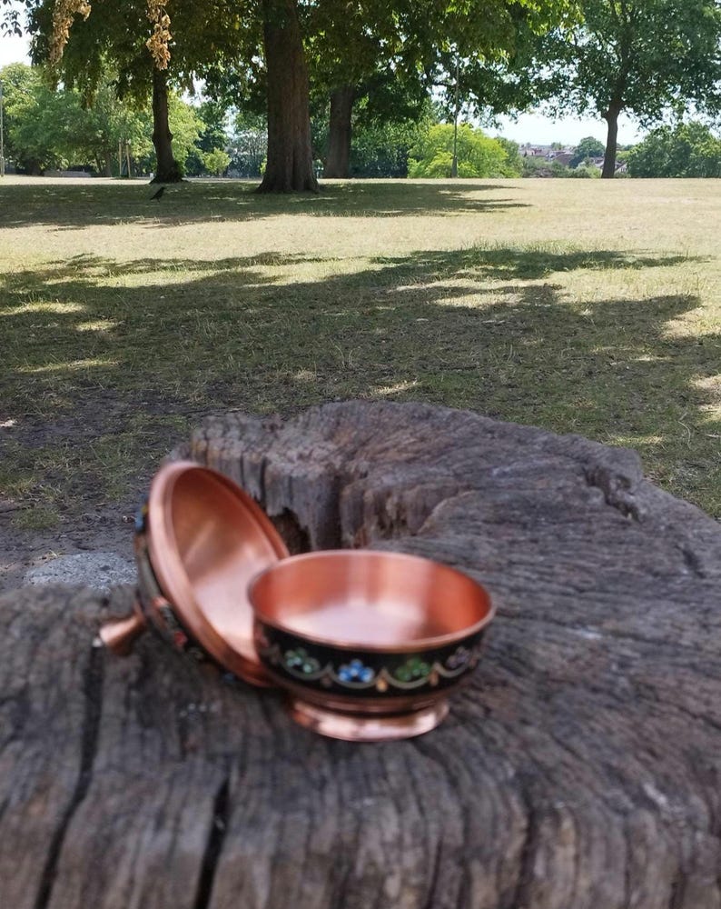 May include: A small, round copper bowl with a matching lid, featuring a black exterior band decorated with a delicate floral pattern in blue, green, and white enamel. The interior of the bowl and lid displays a polished, reflective copper finish.