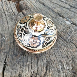 Peut inclure: Un r&eacute;cipient rond en cuivre avec un couvercle dot&eacute; d'un bouton en cuivre poli. Le couvercle est orn&eacute; d'un fond en &eacute;mail cr&egrave;me, de motifs en filigrane noirs et de motifs floraux grav&eacute;s avec une finition en cuivre m&eacute;tallique sur la surface.