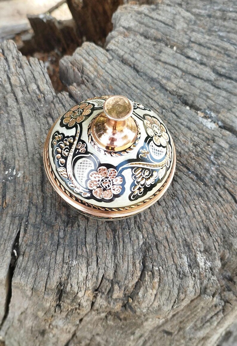 Peut inclure: Un r&eacute;cipient rond en cuivre avec un couvercle dot&eacute; d'un bouton en cuivre poli. Le couvercle est orn&eacute; d'un fond en &eacute;mail cr&egrave;me, de motifs en filigrane noirs et de motifs floraux grav&eacute;s avec une finition en cuivre m&eacute;tallique sur la surface.