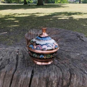 Peut inclure: Un petit r&eacute;cipient rond en cuivre avec un couvercle, dot&eacute; d'une base noire et d'une partie sup&eacute;rieure blanche. La surface est orn&eacute;e de motifs floraux complexes et color&eacute;s dans des tons de bleu, vert et rose, avec un bord et un bouton en cuivre.