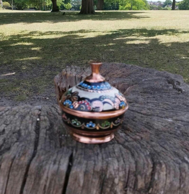 Peut inclure: Un petit r&eacute;cipient rond en cuivre avec un couvercle, dot&eacute; d'une base noire et d'une partie sup&eacute;rieure blanche. La surface est orn&eacute;e de motifs floraux complexes et color&eacute;s dans des tons de bleu, vert et rose, avec un bord et un bouton en cuivre.