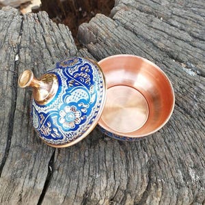 May include: A round copper bowl with a matching lid. The exterior of the lid features a vibrant blue enamel finish with intricate gold and white floral patterns. The lid has a polished copper knob handle, and the interior of the bowl is plain copper.