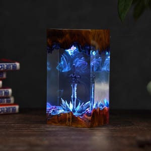 May include: A rectangular resin art piece featuring a detailed sword encased within a clear, glowing blue interior. The top and bottom edges are made of natural wood. The piece is illuminated with blue light, creating a mystical effect.