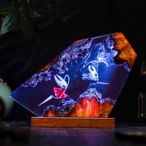 May include: A decorative resin art piece featuring two stylized figures with swords, set against a backdrop of swirling blue and red hues. The artwork is encased in a clear, angled resin block with a wooden base.