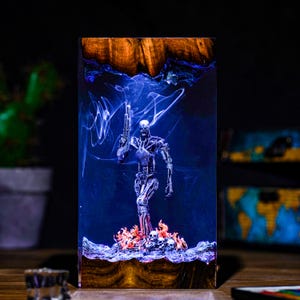 May include: A rectangular resin art piece featuring a metallic robot figure holding a gun, standing on a bed of skulls and flames. The background is a deep blue with wispy white accents. The top and bottom edges are wood.
