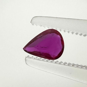 Pode incluir: Uma pedra preciosa em forma de pera, de cor magenta profunda, segurada por uma pinça de metal prateado com bordas serrilhadas. A pedra apresenta múltiplas facetas que refletem a luz, criando uma aparência brilhante sobre um fundo branco.