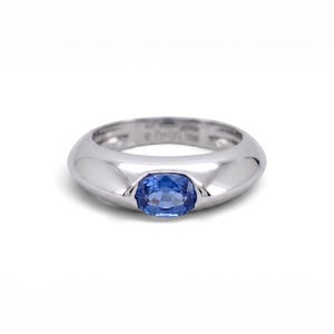 Könnte beinhalten: Ein Ring aus poliertem, silberfarbenem Metall mit einer glatten, abgerundeten Ringschiene und einem einzelnen, oval geschliffenen blauen Saphir, der mittig eingefasst ist. Auf der Innenseite der Schiene sind schwache Gravuren zu erkennen.