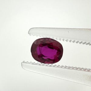 Op de afbeelding: Een diep magenta-kleurige ovale edelsteen met een gefacetteerd oppervlak, vastgehouden tussen de gekartelde metalen punten van een pincet. De steen vertoont een doorschijnende kwaliteit met interne lichtreflecties over de geslepen facetten.