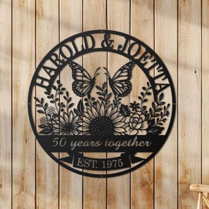May include: Black metal wall art with a circular design featuring the names "HAROLD & JOETTA", butterflies, flowers, and the text "50 years together EST. 1975". The art is mounted on a wooden wall.