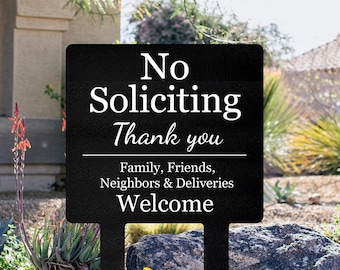 May include: Black metal sign with white text reading "No Soliciting Thank you", and "Family, Friends, Neighbors & Deliveries Welcome". The sign is in a garden with plants and rocks, set against a house.