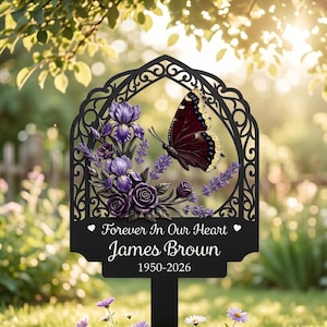 May include: Black metal memorial garden stake with a decorative butterfly and floral design. The stake features the text "Forever In Our Heart James Brown 1950-2026". The butterfly has brown and yellow wings, and the flowers are purple.