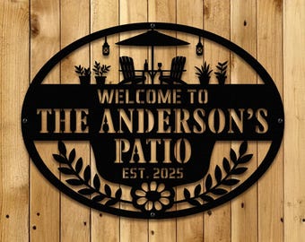 May include: Black metal oval sign with the text "WELCOME TO THE ANDERSON'S PATIO EST. 2025". The sign features a patio scene with chairs, an umbrella, and plants, and is mounted on a wooden background.