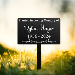 May include: Black metal garden stake with a sign that reads "Planted in Loving Memory of Dylan Hayes 1956 - 2024". The sign is in a field of green grass with yellow flowers.