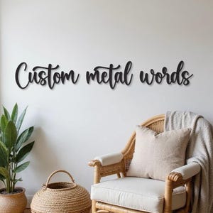 Puede incluir: Arte de pared de metal negro que deletrea "Custom metal words" en una fuente cursiva. Las palabras están montadas en una pared blanca sobre una silla de mimbre con un cojín beige y una cesta tejida.