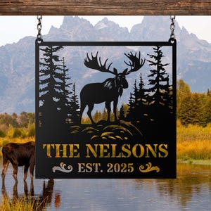 May include: A square black metal sign hanging from two silver chains. The sign features a laser-cut silhouette of a moose standing in a forest of pine trees. Below the moose, the text reads THE NELSONS and EST. 2025 in a gold-colored font.