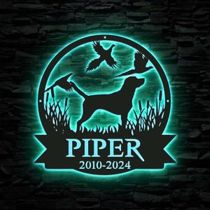 May include: A black metal silhouette of a dog with two pheasants in a circle with the name "PIPER" and the dates "2010-2024" on a black banner below.