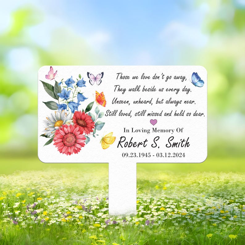 May include: A white memorial garden stake with a floral and butterfly design. The stake includes the text "Those we love don't go away... In Loving Memory Of Robert S. Smith 09.23.1945 - 03.12.2024."