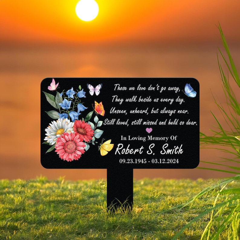 May include: A black memorial garden stake with a rectangular sign featuring a floral design and butterflies. The sign includes the text "Those we love don't go away... In Loving Memory Of Robert S. Smith 09.23.1945 - 03.12.2024."