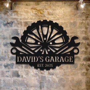 May include: Black metal garage sign with the text "DAVID'S GARAGE" and "EST. 2025". The sign features a gear, two wrenches, and a wheel design. The sign is mounted on a brick wall.