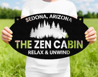 May include: Black metal sign with the text "SEDONA, ARIZONA" and "THE ZEN CABIN RELAX & UNWIND". The sign features a cut-out forest silhouette. The sign is oval-shaped with two holes at the top for hanging.