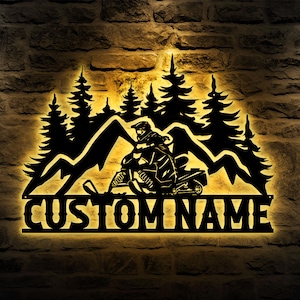 May include: Black metal wall art featuring a snowmobiler riding through a mountain scene with pine trees. The words "CUSTOM NAME" are displayed below the scene. The art is illuminated with a warm, yellow glow, creating a striking contrast against the dark metal.