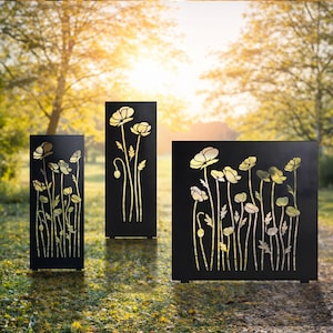 Puede incluir: Tres pantallas de jardín de metal negro con un diseño floral. Las pantallas son rectangulares y presentan imágenes recortadas de flores y tallos en color dorado. Las pantallas están colocadas al aire libre en una zona de césped.
