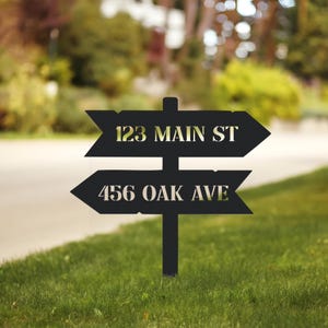 May include: Black metal address sign with two directional arrows. The top arrow reads "123 MAIN ST" and the bottom arrow reads "456 OAK AVE". The sign is mounted on a post and set in a grassy yard.