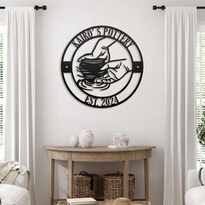 May include: Black metal wall art featuring a pottery design with the text "Kairo's Pottery" and "EST. 2024". The artwork depicts hands shaping a clay pot. The piece is circular and mounted on a white wall.