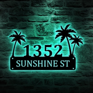 Puede incluir: Placa de número de casa de metal negro con siluetas de palmeras y la dirección "1352 SUNSHINE ST". La placa está iluminada con una luz turquesa brillante.