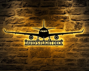 Puede incluir: Una silueta de avión de metal negro con el texto "DAVID'S FLIGHT DECK" debajo. La silueta está iluminada desde atrás, proyectando un brillo cálido en una pared de ladrillo.