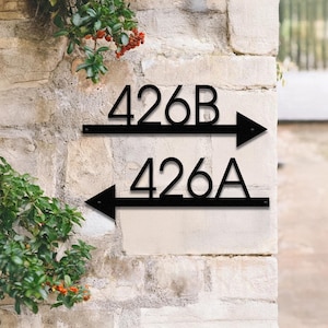 May include: Black metal address signs with the numbers 426A and 426B. The numbers are in a bold, sans-serif font. Each sign has an arrow pointing left or right. The signs are mounted on a light-colored stone wall, with greenery in the background.