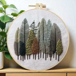 May include: An embroidered forest scene in a wooden hoop. The artwork features a variety of textured trees in shades of green, brown, and gray, with dark vertical lines representing tree trunks. The background is a light-colored fabric.