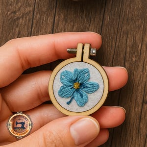 May include: A small embroidery hoop with a light wood frame. The hoop features a blue flower design with a yellow centre on a piece of fabric. The hoop is held in a person's hand. The embroidery is a miniature craft item.