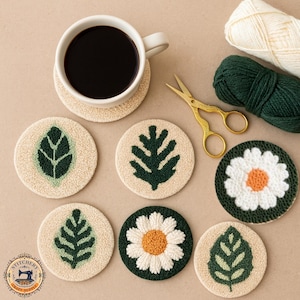 May include: Six circular punch needle coasters displaying botanical and floral patterns in cream, dark green, and orange. One coaster holds a white ceramic mug containing black coffee. Gold-coloured metal scissors and two skeins of yarn are included.