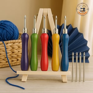 May include: A set of five punch needle tools with colorful wooden handles in purple, green, red, yellow, and blue. The tools are displayed on a small wooden easel, with additional needles and a ball of blue yarn in the background. Craft supply.