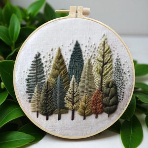May include: A hand-stitched embroidery hoop art piece featuring a forest scene. The artwork showcases a variety of detailed trees in shades of green, brown, and gold, set against a white fabric background. The embroidery is framed in a natural wood hoop.