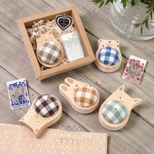 May include: A selection of wooden pin cushions in animal shapes, such as cats and bears, each with a checkered fabric cushion. The set includes a gift box with a heart-shaped tag that reads "For the love of sewing."