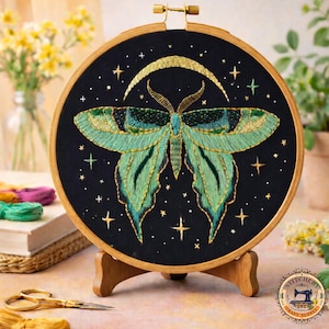 May include: An embroidered moth design on a black background, set in a wooden hoop. The moth is detailed with green and gold thread, with a crescent moon and stars. The hoop is displayed on a wooden stand, with embroidery supplies nearby.