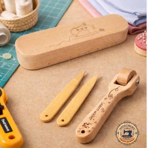 May include: A collection of wooden sewing tools, including a seam roller, two point turners, and a ruler box with a sewing machine design. The tools are light brown and sit on a tan surface. A rotary cutter and other sewing supplies are also visible.