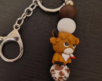 May include: A keychain with a silver ring and chain. The keychain features a brown and white spotted bead, a cartoon Highland cow figure, a white bead, and a brown bead. The cow has a red tongue sticking out.
