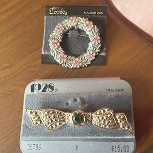 May include: Two vintage brooches on display. The top brooch is a wreath design with pink and green enamel flowers. The bottom brooch is a bow shape with a green center stone and gold-tone metal. Both are on gray velvet cards.