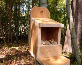 Puede incluir: Un comedero de pájaros de madera con una pequeña abertura para que los pájaros puedan acceder a los cacahuetes del interior. El comedero está montado en el tronco de un árbol y tiene una pequeña plataforma debajo de la abertura.