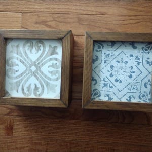 May include: Two square framed wall decor pieces with a white background and intricate patterns. The left frame has a white and gray pattern, while the right frame has a blue and white pattern.