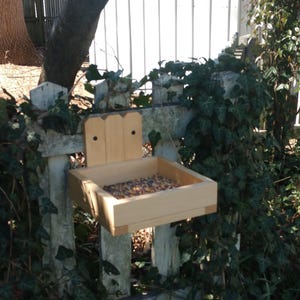 May include: A wooden bird feeder with a tray filled with birdseed, mounted on a white picket fence with green ivy growing around it.