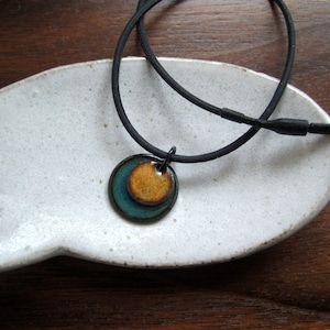 May include: A black cord necklace with a round pendant featuring concentric circles of teal, brown, and gold enamel.