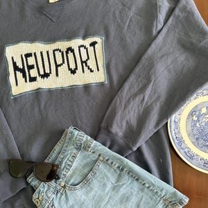 Puede incluir: Una sudadera de cuello redondo color azul pizarra con un parche rectangular de punto que muestra la palabra NEWPORT en letras negras sobre un fondo crema. Incluye pantalones cortos de mezclilla azul claro, gafas de sol negras y un plato.