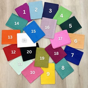 Pode incluir: Uma coleção de vinte e uma peças de tecido quadradas em várias cores, dispostas em um padrão circular. As cores incluem roxo, azul, marinho, verde, verde-azulado, laranja, vermelho, preto, rosa, amarelo e branco. Cada peça está numerada.