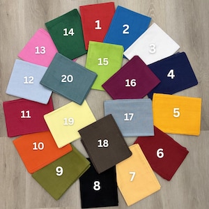 May include: A selection of twenty folded square fabric napkins in a variety of colours, such as red, blue, white, and green. Each napkin is numbered from 1 to 20, arranged in a circular pattern on a light wood surface.