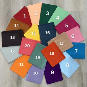 May include: A collection of twenty folded square fabric napkins in various colors, including red, orange, green, blue, and purple. Each napkin is numbered from 1 to 20, arranged in a circular pattern on a light wood surface.