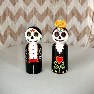 May include: A pair of Day of the Dead-themed wooden peg dolls. The dolls are painted in black, white, and red with skull faces and floral details. The female doll has a yellow flower on her head.